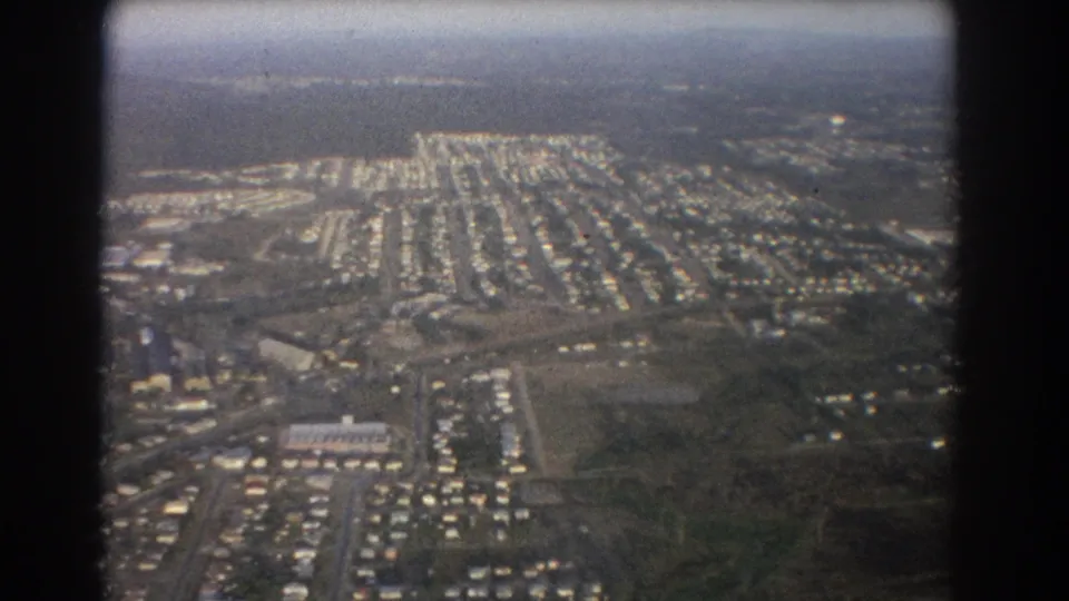 Brisbane 1967 aerial frame with suburban homes, roads, open lots, and a hazy horizon captured on 8mm film.