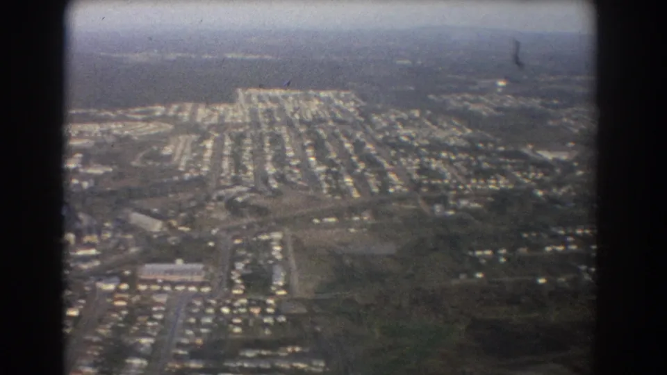 1967 Brisbane aerial home-movie frame showing dense low-rise suburban streets beyond dark airplane-window borders.