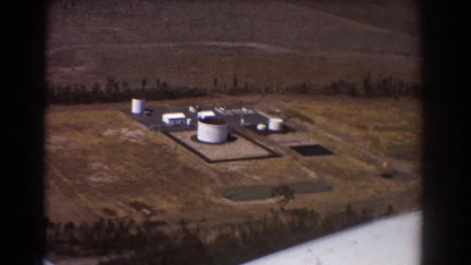 1967 Brisbane-area aerial frame showing a rural industrial site with tanks, small structures, fields, and an aircraft wing edge.