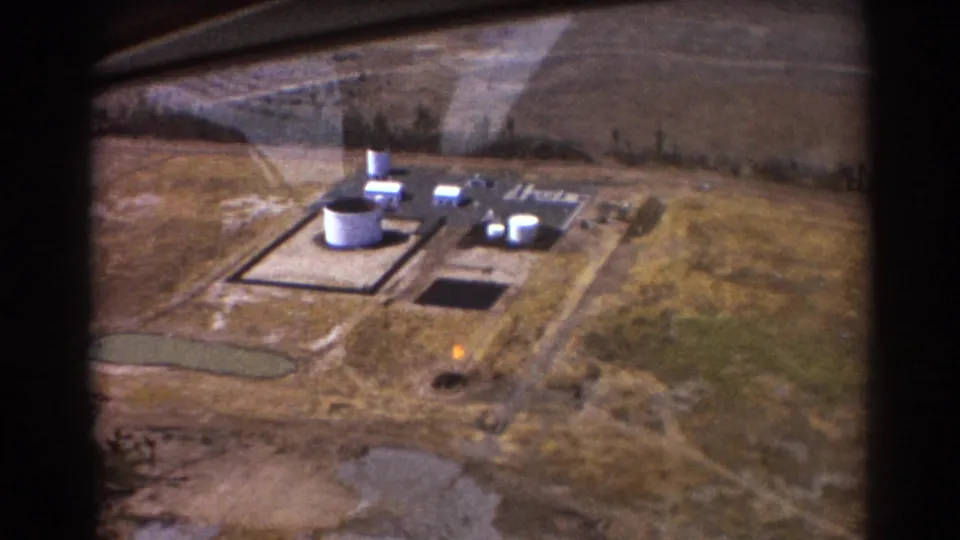 1967 Brisbane aerial home-movie frame with industrial tanks, rectangular structures, small ponds and dry surrounding land.