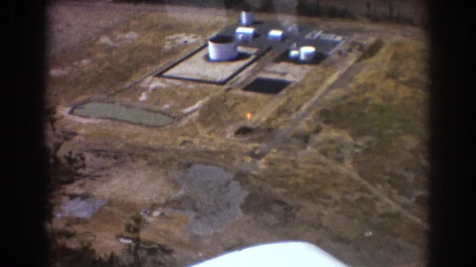 Aerial 8mm frame of the Brisbane industrial facility, showing white tanks, low buildings, service roads, ponds and aircraft-edge framing.