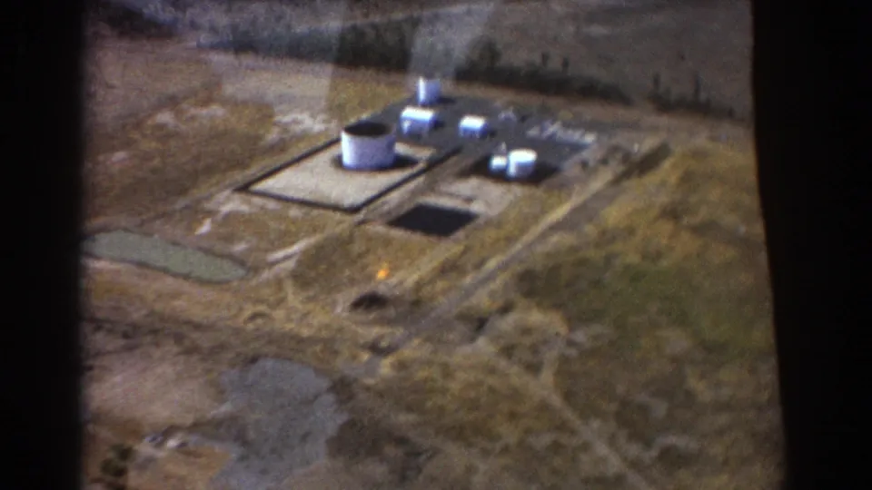 1967 Brisbane aerial 8mm frame showing an industrial plant with white cylindrical tanks, paved pads, dirt tracks, and dry surrounding land.