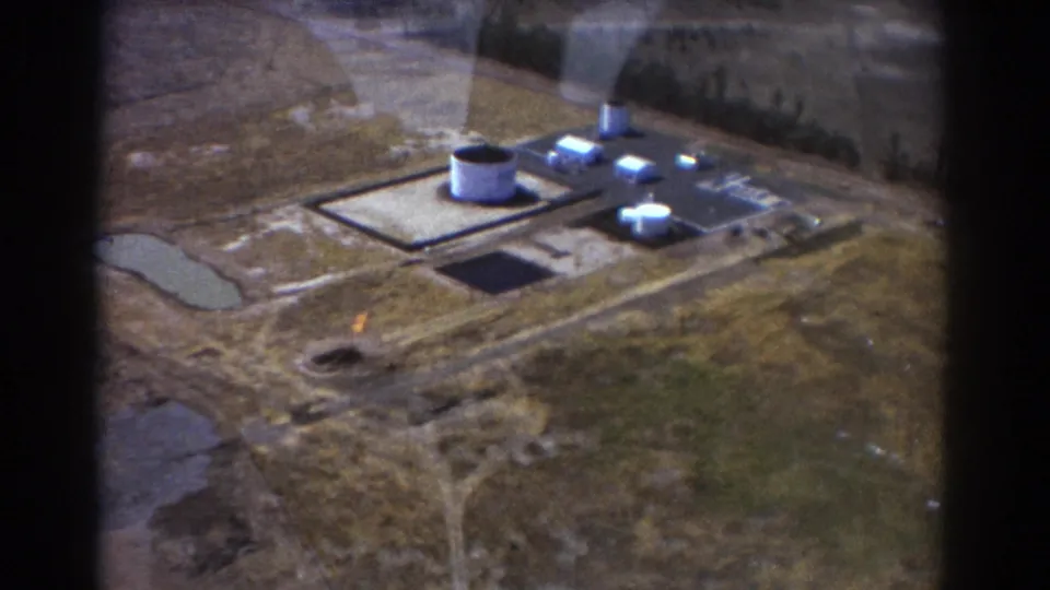 1967 Brisbane 8mm aerial view showing an industrial plant, white tank structures, rectangular paved areas, dry land, and a small visible flame.