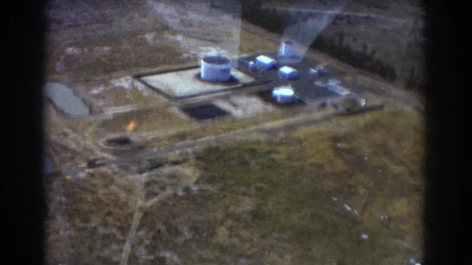 1967 Brisbane aerial frame of a remote industrial complex, with white tanks, low buildings, dark paved areas, and aircraft-window edges in view.