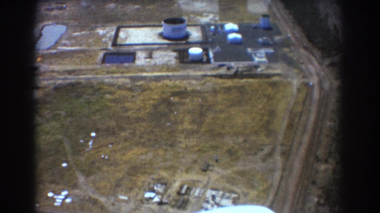Industrial site seen from above, with a large round tower, paved areas, fields, and rail line.