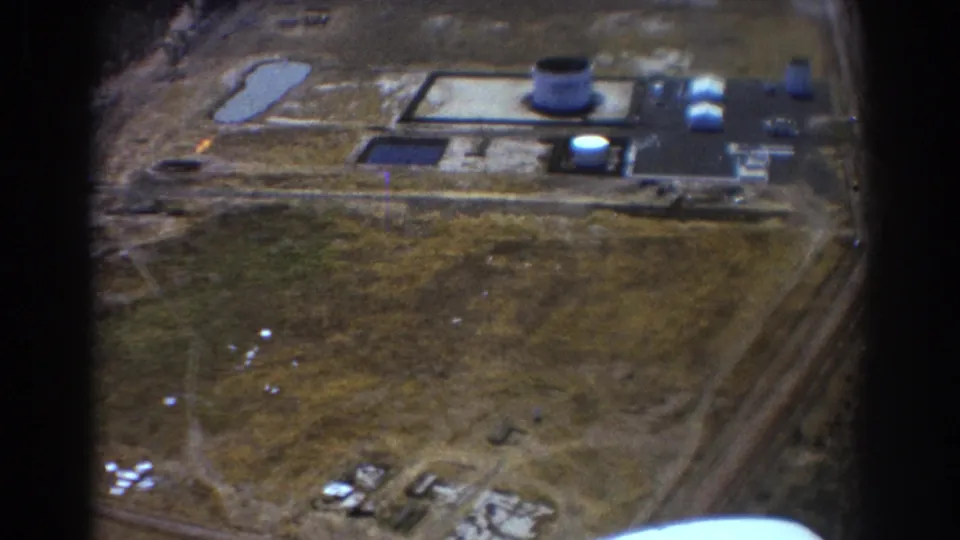 1967 home-movie aerial frame showing an industrial complex from above, with tanks and roads partly interrupted by dark airplane-window or film-edge shadows.