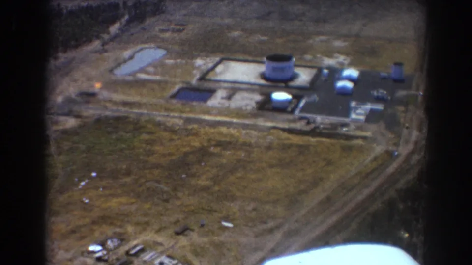 Brisbane 1967 airplane-window view showing the industrial site receding below, with tanks, low structures, fields, rail line, and a bright window edge.