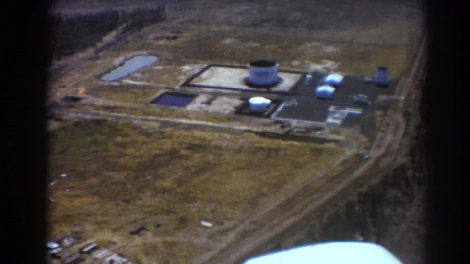 1967 Brisbane aerial 8mm frame showing an industrial complex with a large cylindrical tank, smaller tanks, buildings, roads, ponds, and open ground.