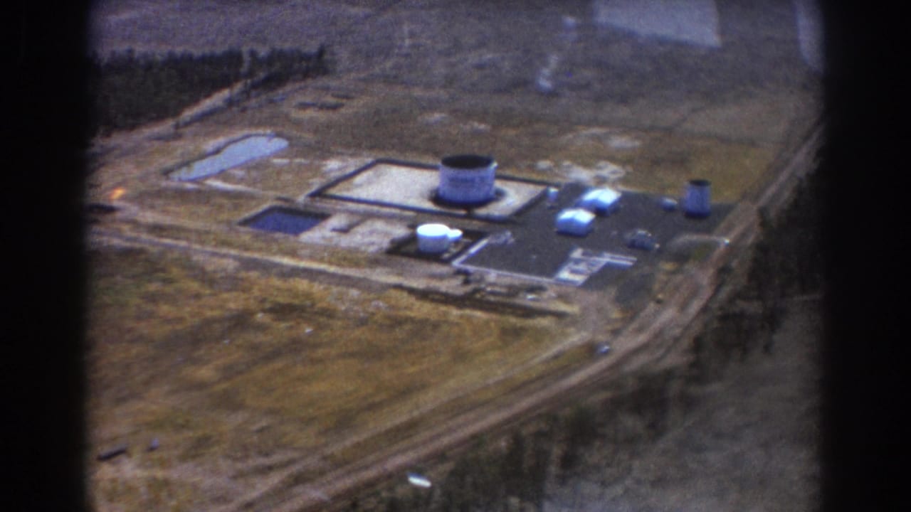 Industrial site seen from above with central tank, low buildings, roads, ponds, and sparse trees.