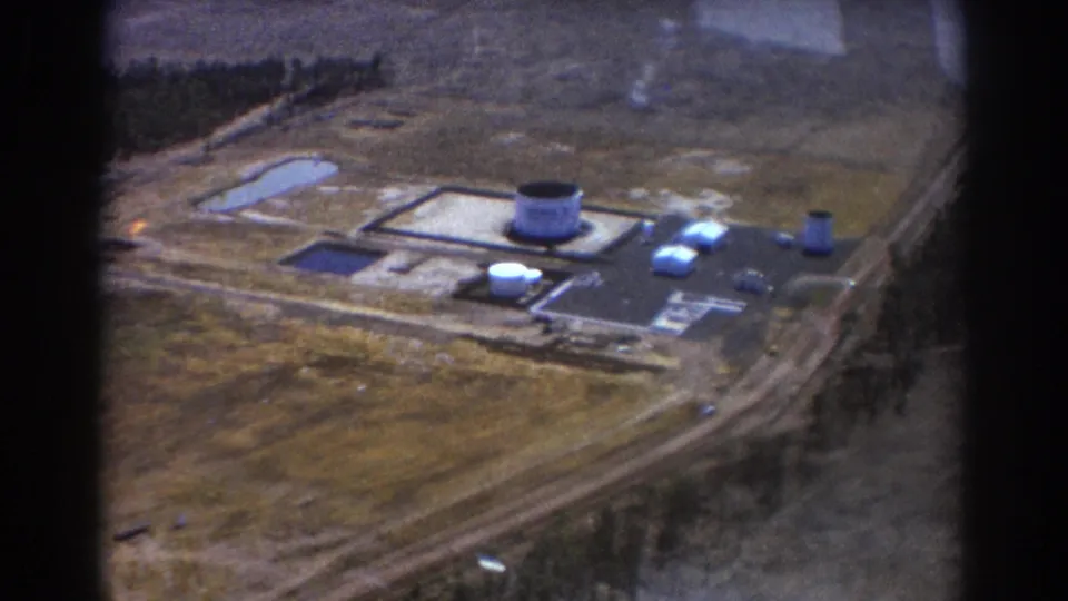 1967 Brisbane aerial frame with the industrial complex centered, showing tanks, low rectangular structures, access roads, ponds, and surrounding open terrain.