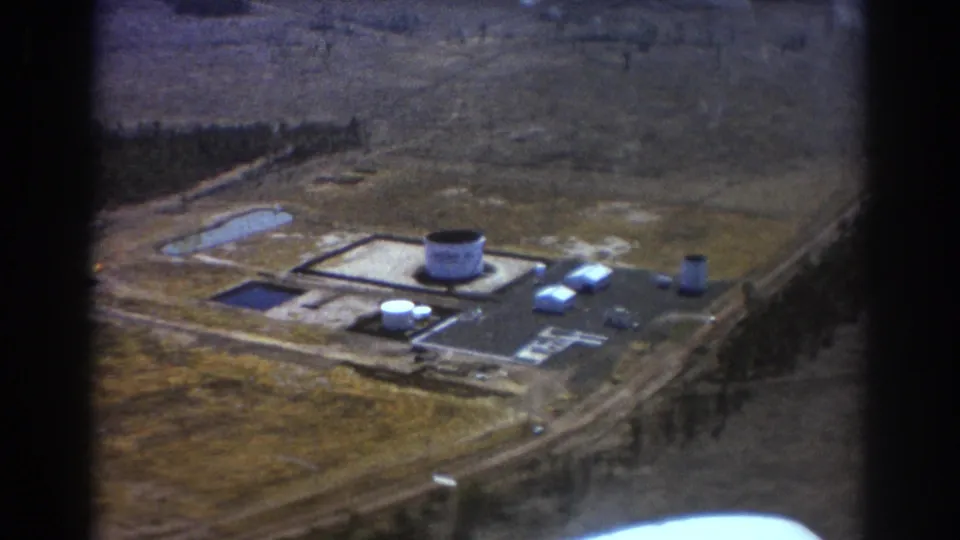 1967 Brisbane aerial 8mm view of an industrial complex from a slightly lower angle, with tanks, buildings, dirt roads, fields, and trees visible.