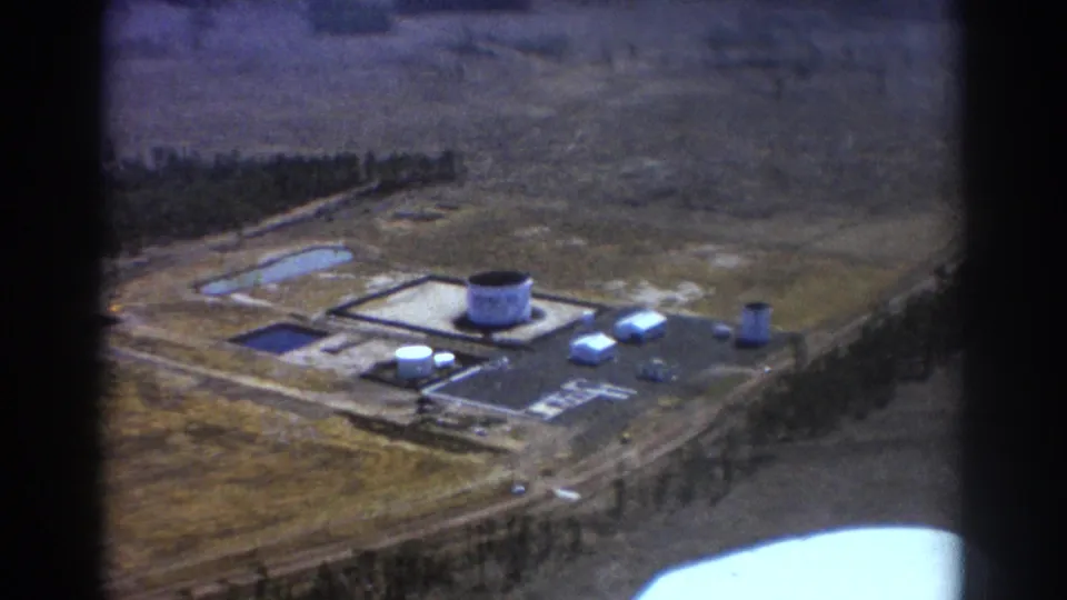1967 Brisbane aerial frame showing the industrial site receding below, with tanks, buildings, roadways, trees, and open surrounding land.