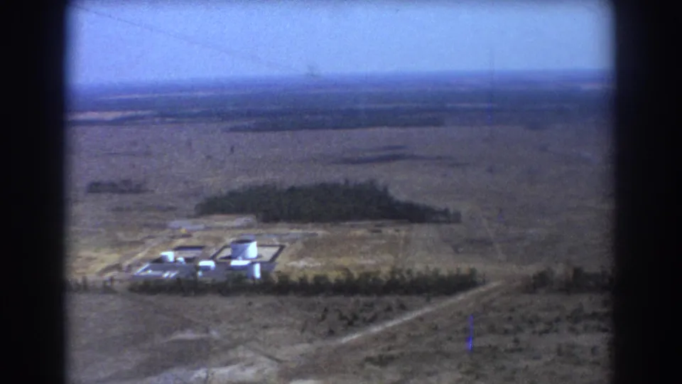 1967 Brisbane-area aerial view of open plains with white cylindrical tanks, low buildings, tree lines, and a distant horizon.