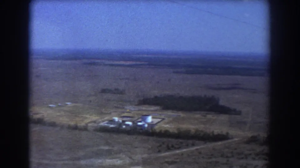 1967 aerial home-movie view near Brisbane with the industrial site lower in frame, surrounded by plains and a dark tree belt.