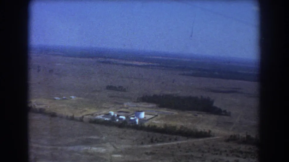 Brisbane 1967 aerial frame with white tanks, low structures, a tree line, and broad plains receding toward the horizon.