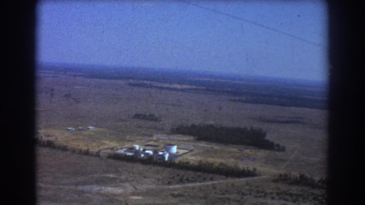 Aerial view over dry plains with a small airstrip area and scattered trees below.