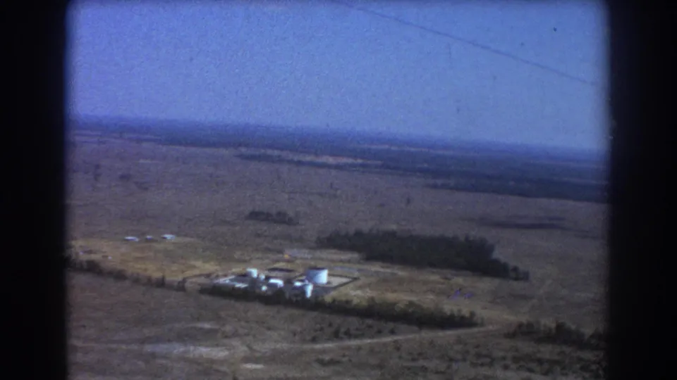 1967 8mm aerial view near Brisbane, Australia, showing dry open land, tree lines, and a small airstrip area seen from an airplane.