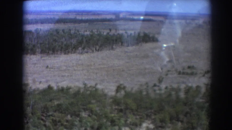 Low 8mm aerial view near Brisbane showing dry fields, scattered trees, and distant runway area during the aircraft approach.