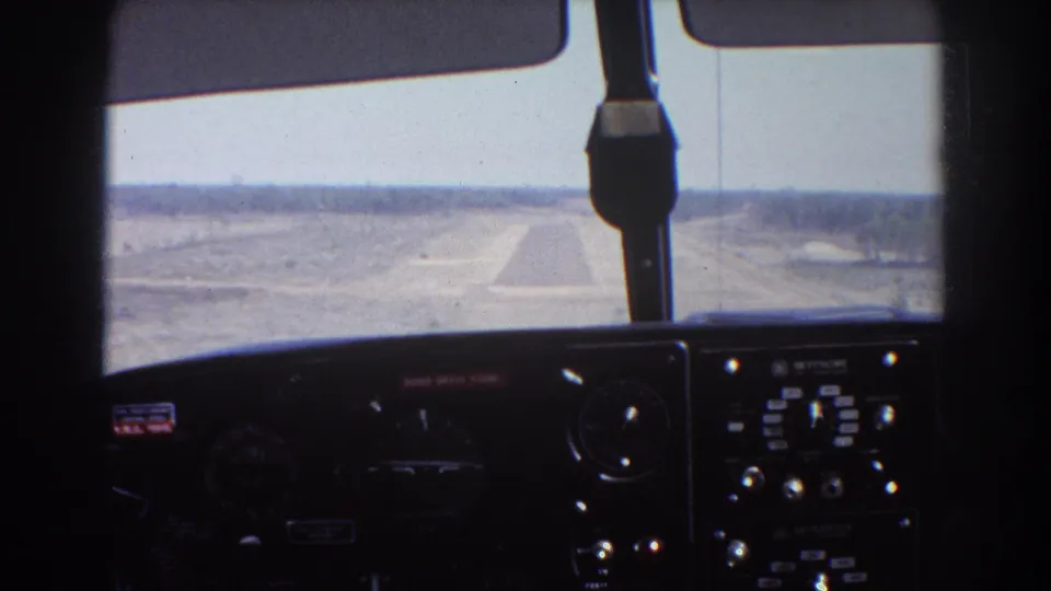 Brisbane-area 1967 cockpit landing view with aircraft instruments in shadow and a remote runway ahead over dry ground.
