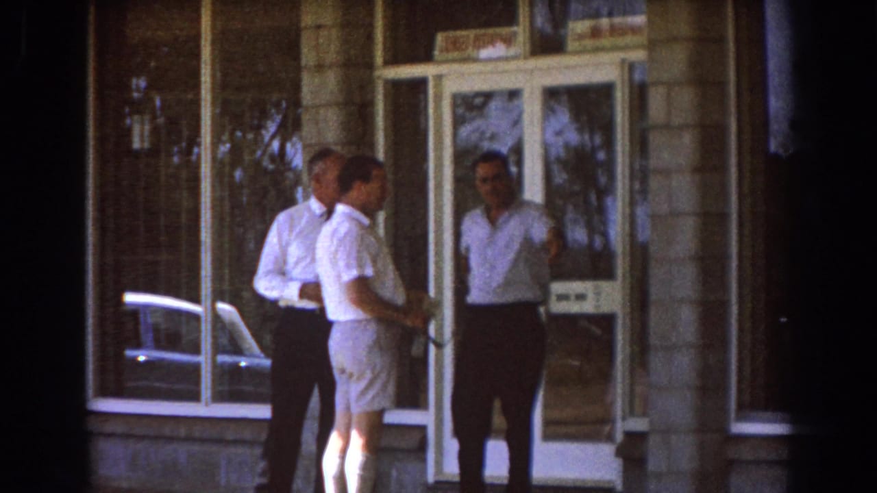 Three adult men in light shirts stand near a doorway outside a Brisbane building in 1967 home-movie footage.