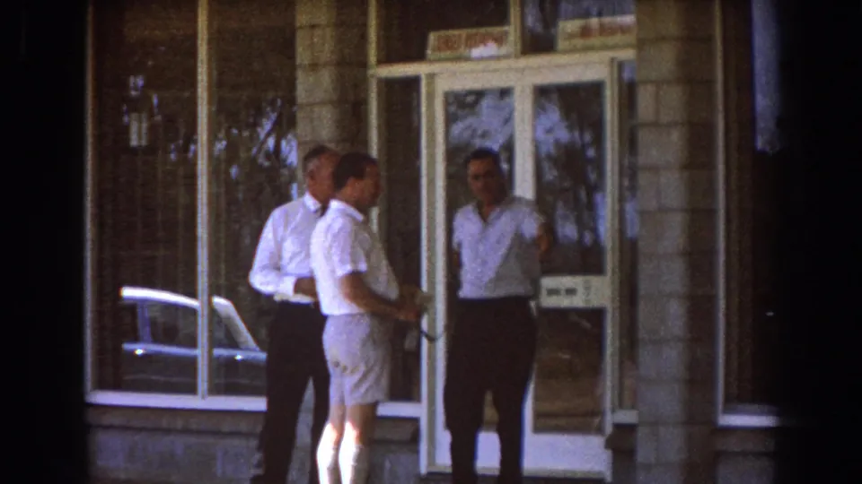 Three adult men in light shirts stand near a doorway outside a Brisbane building in 1967 home-movie footage.
