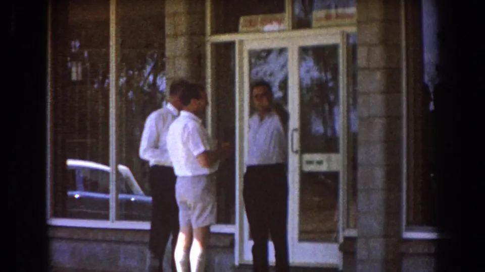 Two men on the sidewalk speak with a third man at the glass entrance of a Brisbane building.