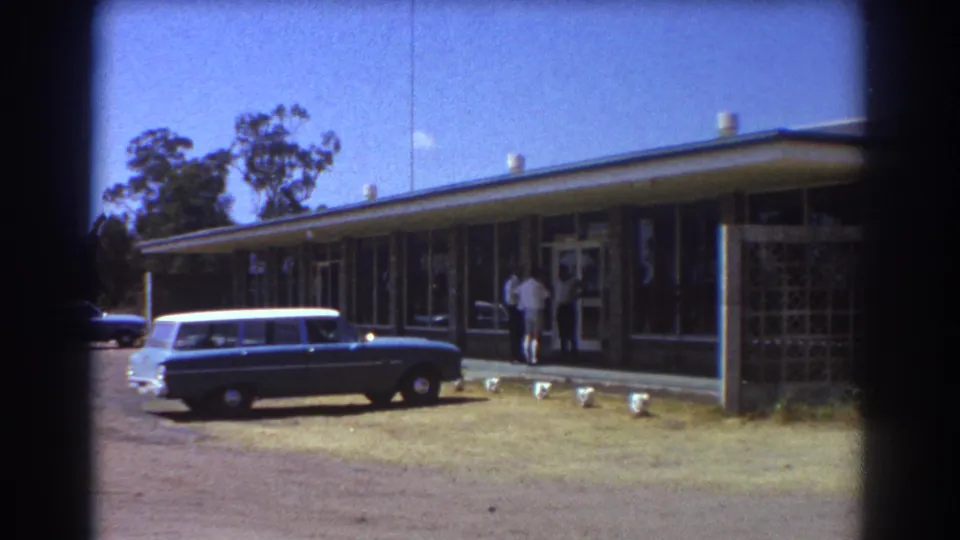A blue station wagon with a white roof is parked near the building where the men stand in Brisbane, 1967.