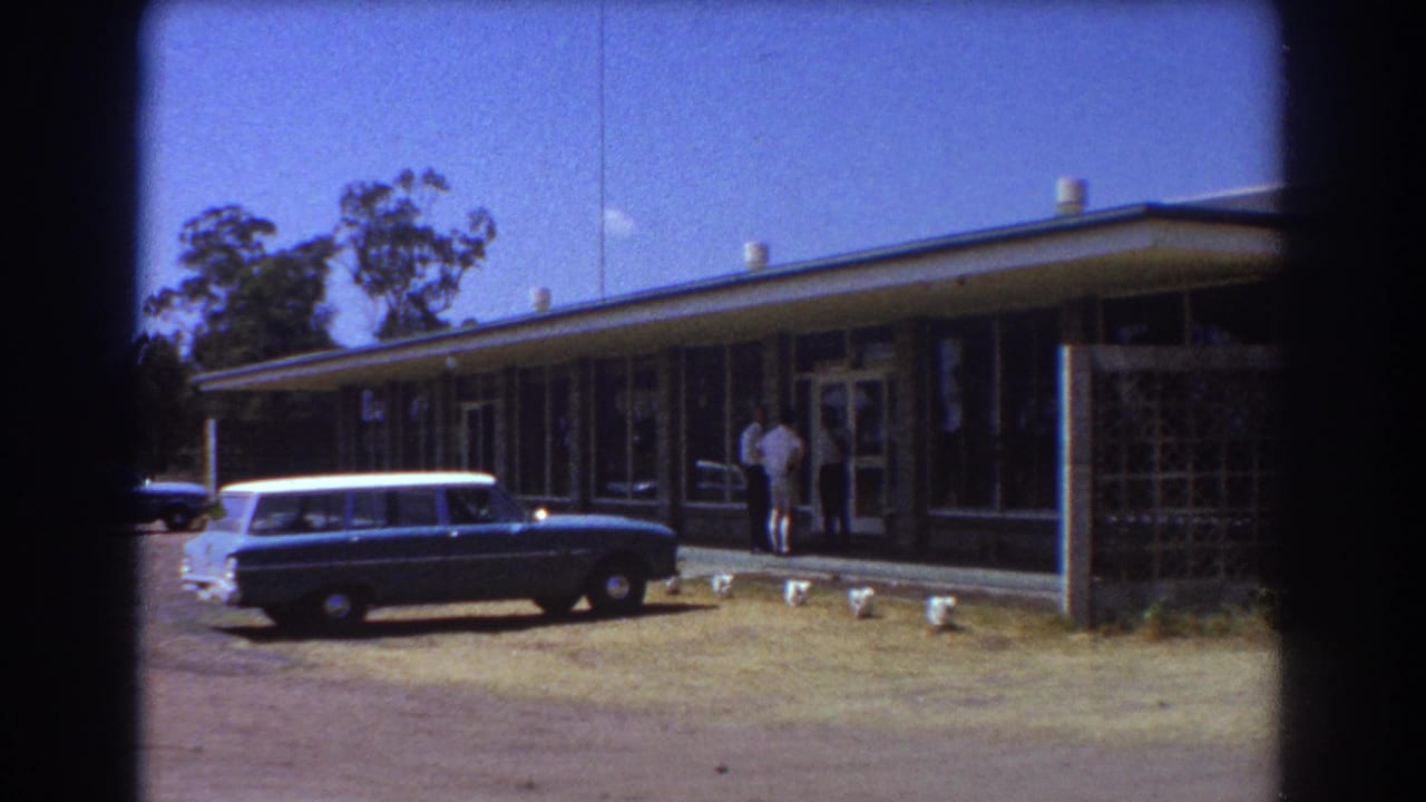 Blue station wagon parked outside a low modernist building on a sunny lawn.