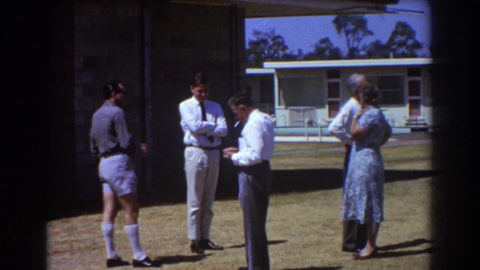 1967 Brisbane frame of four adults in 1960s clothing gathered on a lawn outside a modernist building.
