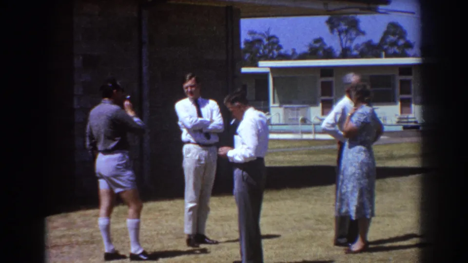 1967 Brisbane home-movie frame showing adults in conversation on the lawn, framed by dark film edges.