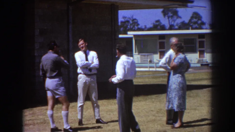 1967 Brisbane frame of adults in 1960s attire standing outside a low building during a sunny conversation.