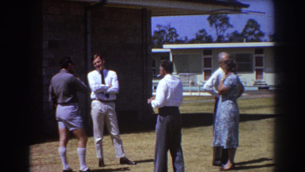 1967 Brisbane home-movie frame of adults conversing outdoors on a lawn beside a low modern house.