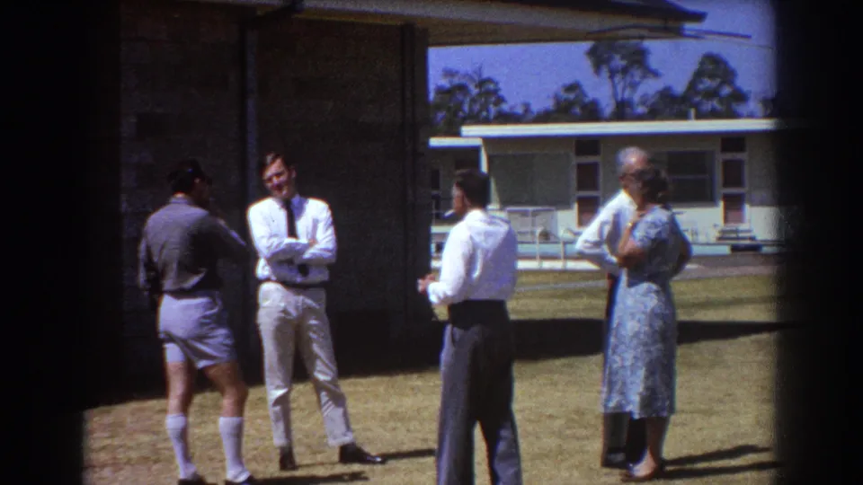 1967 Brisbane home-movie frame of adults conversing outdoors on a lawn beside a low modern house.