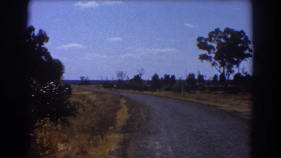 1967 Brisbane home-movie view of a quiet road bordered by dry grass, scrub, and distant trees.