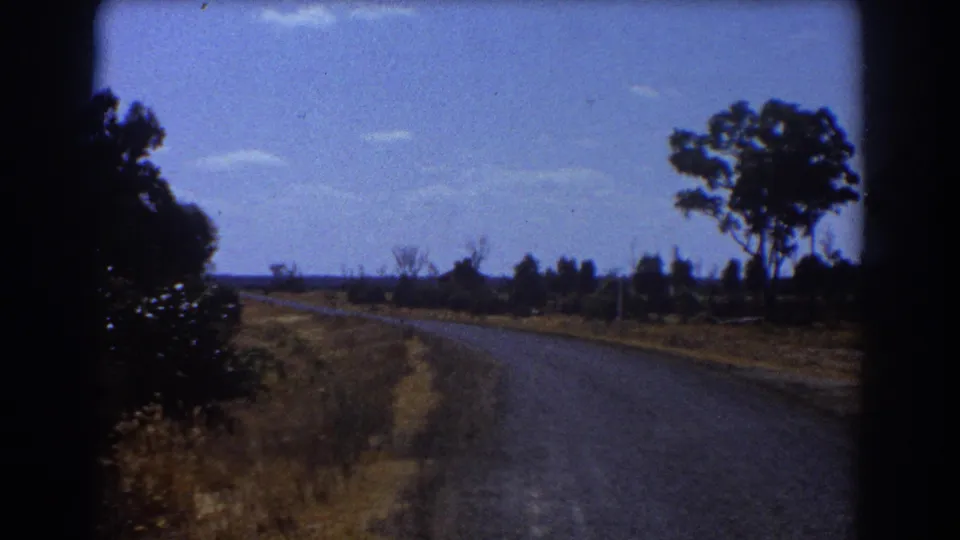 1967 Brisbane home-movie frame showing a quiet road edge, dry grass, tree line, and open blue sky.