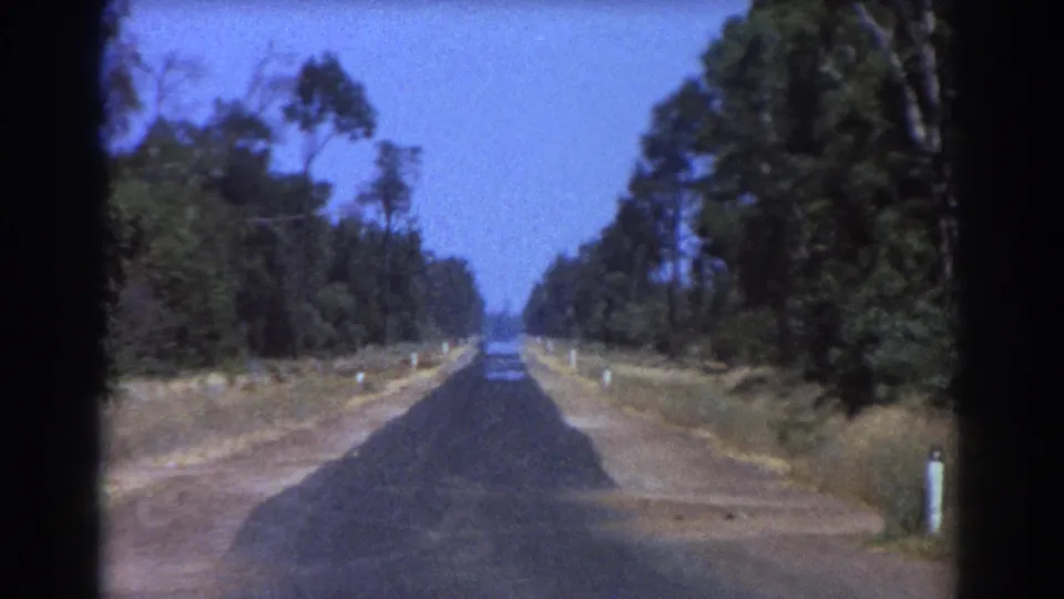 1967 Brisbane-area 8mm frame showing an empty rural road, tree cover on both sides, dry verge, and a low horizon.