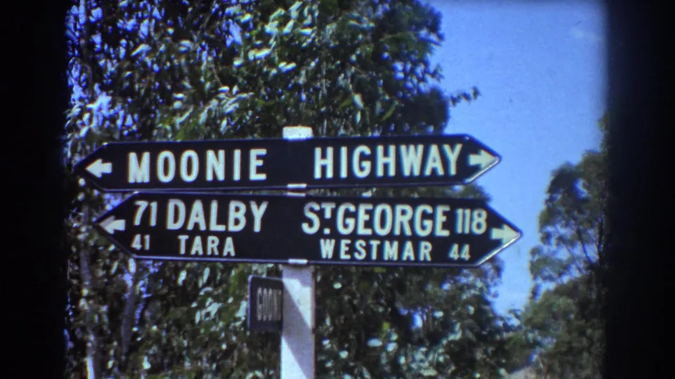 1967 Brisbane-area 8mm frame showing a black directional sign for Moonie, Dalby, St George, Tara, and Westmar beside trees.