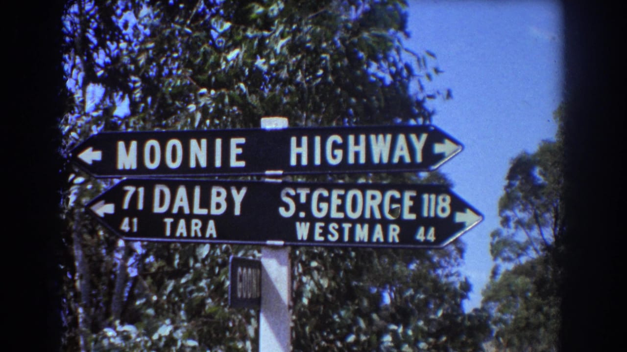 1967 Australian 8mm frame showing a black roadside direction sign for Moonie Highway and regional destinations against trees and blue sky.