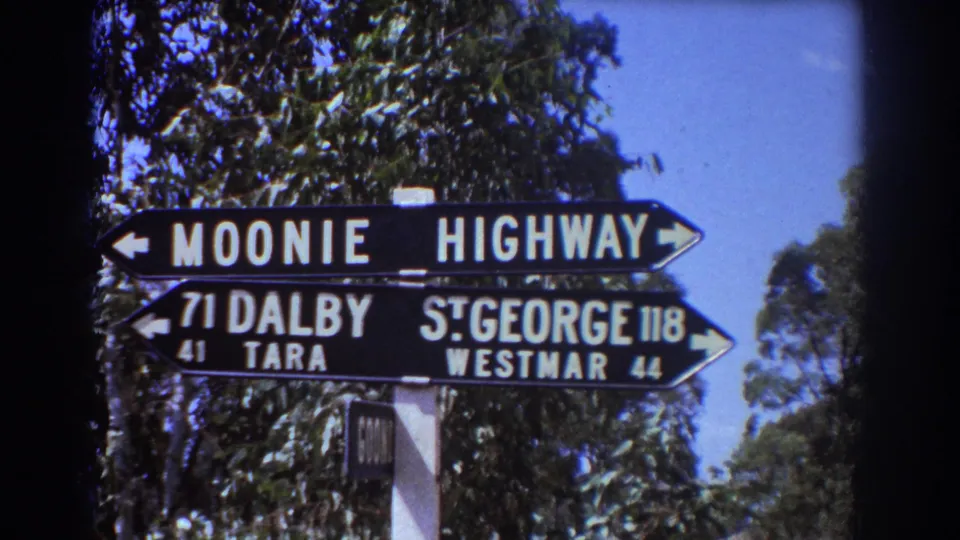 1967 Brisbane-area travel footage frame with a black highway direction sign listing Dalby, Tara, St. George, and Westmar.