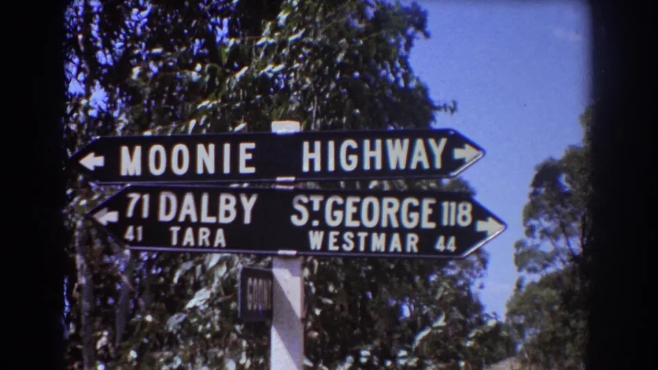 1967 Australian home-movie frame showing the regional road sign and its white support post amid roadside trees.