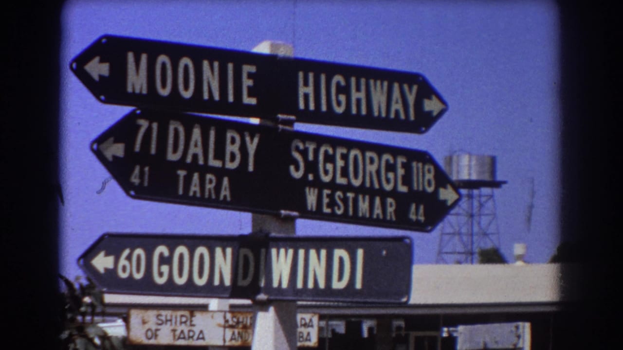 Stacked black-and-white directional signs in supplied 1967 Australian travel footage, with a water tower visible beyond the signpost.