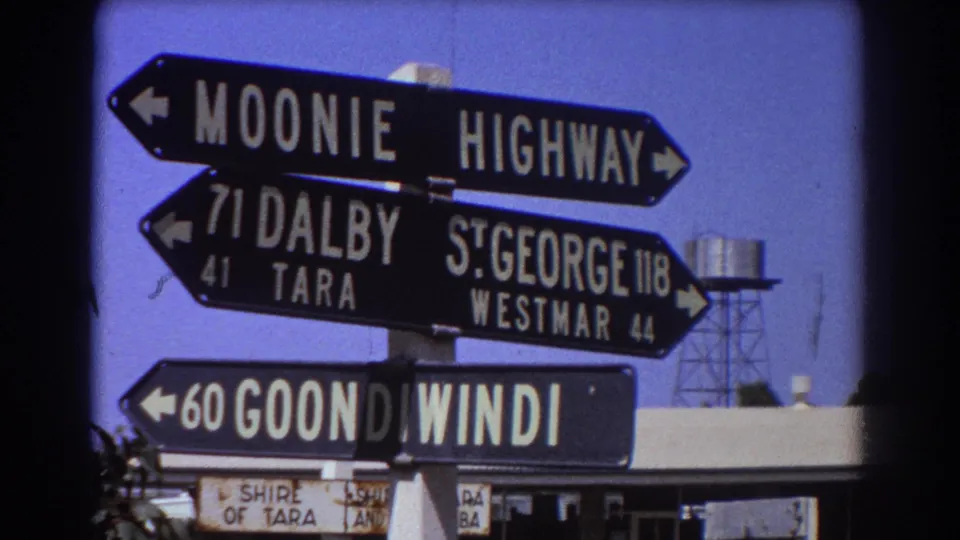 Stacked black-and-white directional signs in supplied 1967 Australian travel footage, with a water tower visible beyond the signpost.