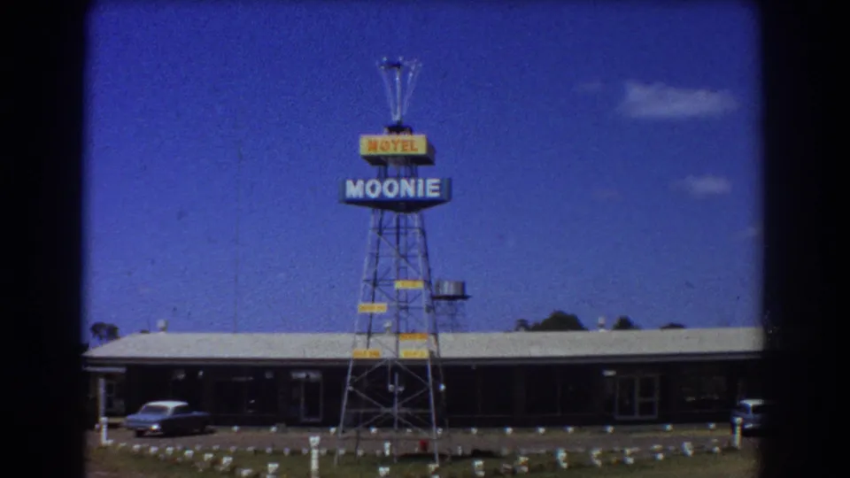 1967 8mm frame showing the Moonie Motel tower sign, low motor-lodge building, blue sky, and parked cars in Australia.