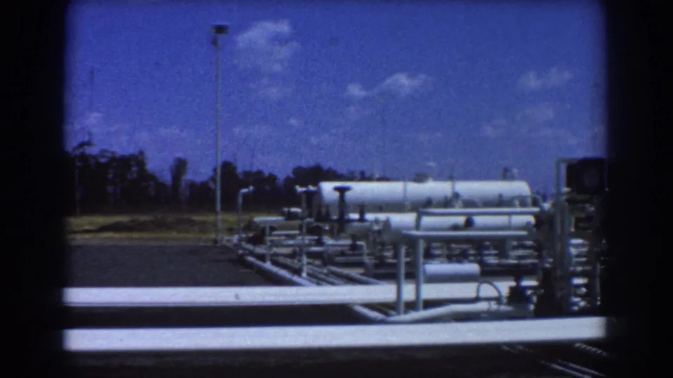 1967 Brisbane footage showing oil yard tanks and low pipework with an open area, trees, and bright sky beyond.
