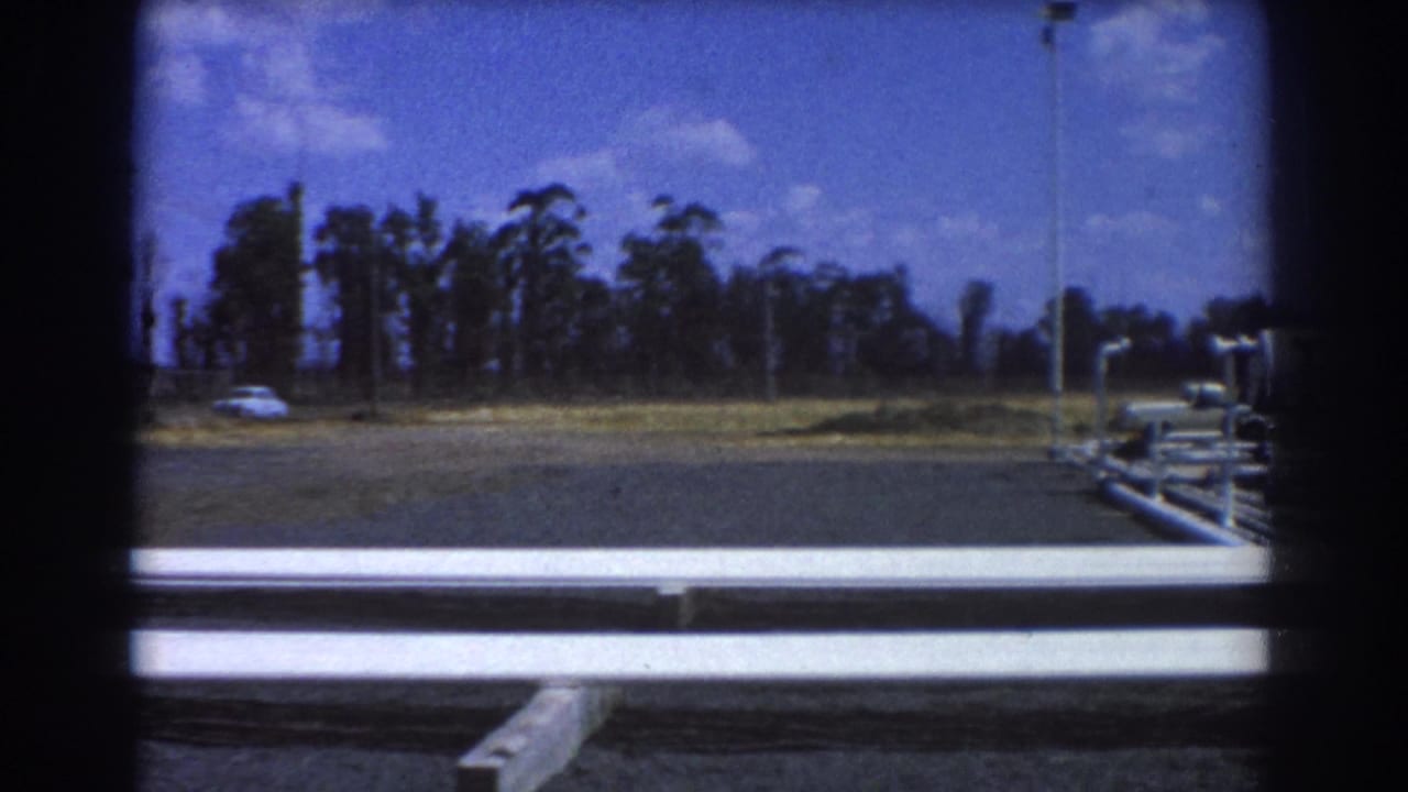 1967 Brisbane roadside industrial view with white pipework, an open paved lot, distant car, trees, and light poles.