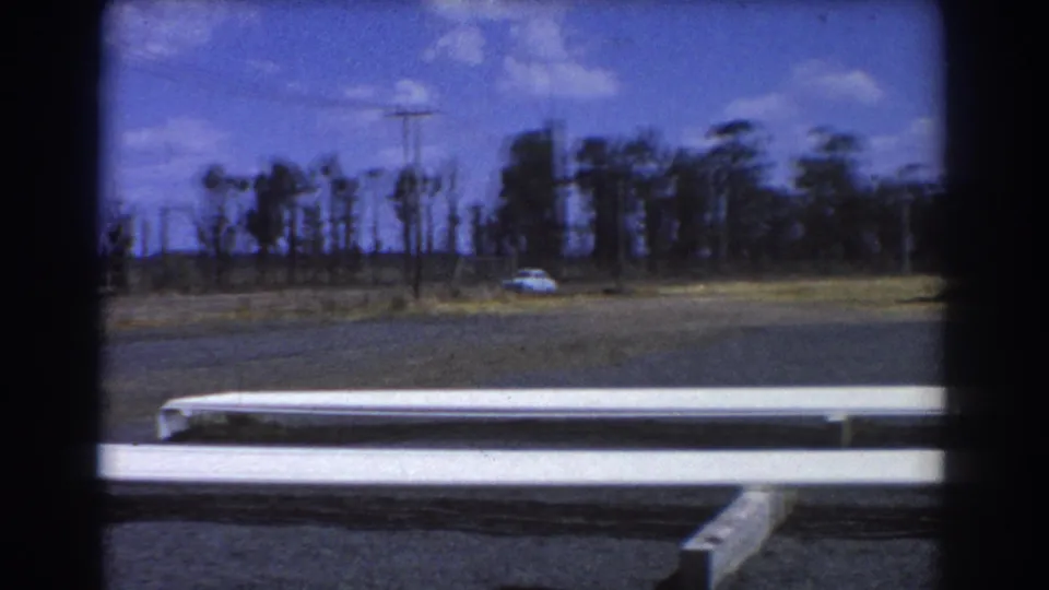 1967 Brisbane 8mm frame with foreground white pipes, open asphalt, a distant white car, utility poles, trees, and clouds.
