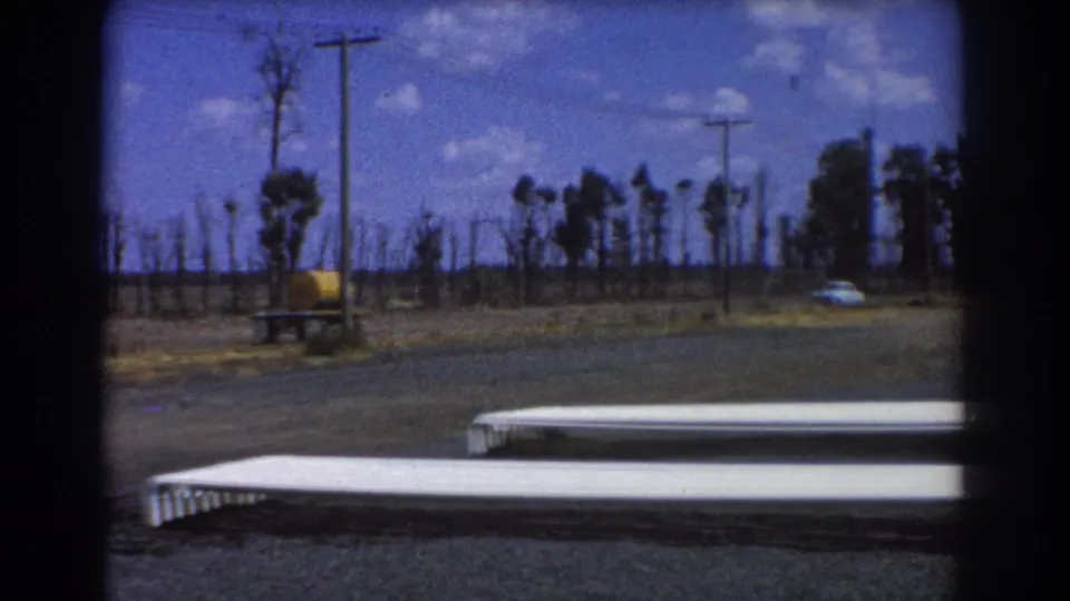1967 Brisbane industrial roadside frame with white pipes, asphalt, utility poles, sparse trees, grass, and a distant white car.