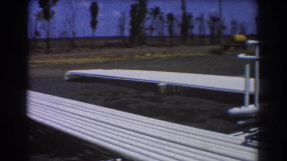 1967 Brisbane 8mm frame shifted away from the machinery toward a paved yard, white railings, and distant palm trees.