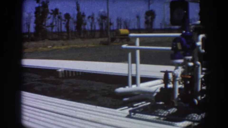 1967 Brisbane 8mm frame showing sunlit white industrial pipework, blue valve fittings, a paved walkway, and distant palm trees.