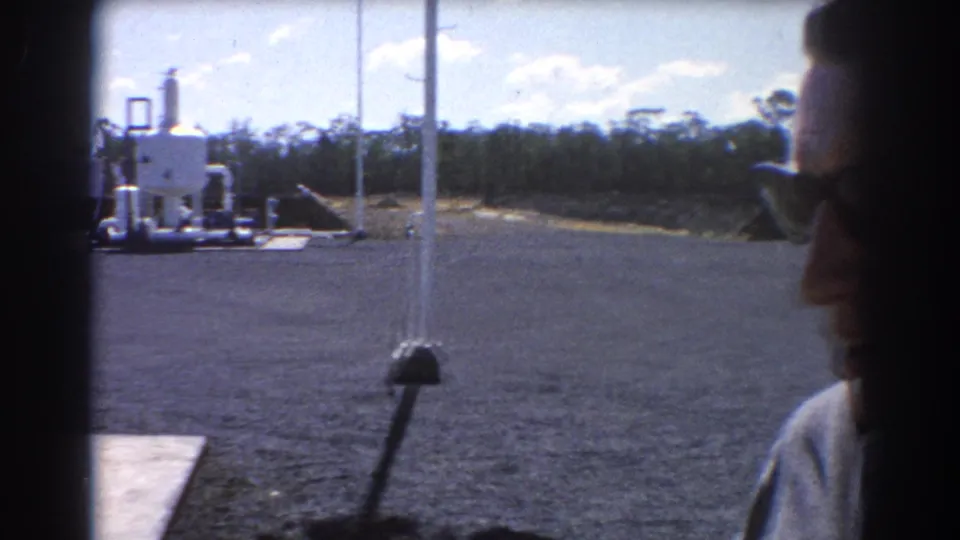 1967 Brisbane home-movie frame showing a gravel industrial yard, white oil equipment, blue sky, and a man in sunglasses partly entering the shot.